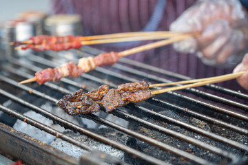 Beef meat on wooden skewer sticks are grilling on the metal grating with smoking from charcoal. Asian traditional streed food object, close-up.