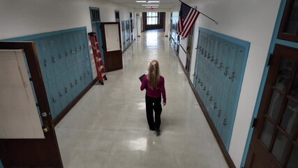 A middle-aged teacher walks away from camera in and empty school hallway holding a tablet. The walls are lined with lockers and large windows let in natural light.