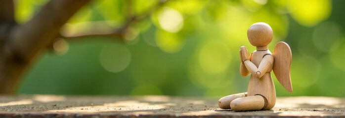 Angel wood man figurine is holding its hands together in a prayer posit with green nature bokeh background.