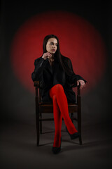 Beautiful girl in a black oversized blazer and red tights posing on the chair on a dark background.