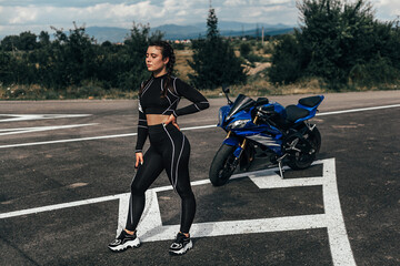 Naklejka na ściany i meble The girl stands in a pose with one hand on her hip and the other hand down. There is a blue sports motorcycle in the background. In the distance you can see trees and mountains under a cloudy sky.