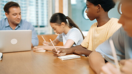 Young smart teenager writing and talking about topic at classroom. Diverse highschool student taking a note in other notebook while sitting with multicultural friends. Creative education. Edification.