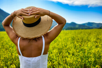 Beautiful woman draped in white dress and straw hat standing in mustard field with scenic view