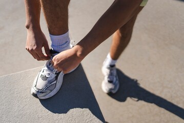 A close-up of hands tying shoelaces on white athletic sneakers while one foot rests on a concrete ledge with sunlight casting shadows on the ground