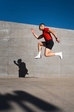 A man in a red shirt and black shorts leaps mid-air, casting a shadow against a gray concrete wall outdoors under a vivid blue sky