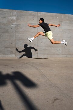 A man wearing yellow shorts and a black shirt leaps dynamically in the air, casting a shadow on a gray concrete wall under a bright blue sky