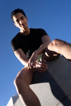 A low-angle view of a man in a black shirt and yellow shorts sitting on a ledge under a vivid blue sky with sunlight creating shadows on the surface