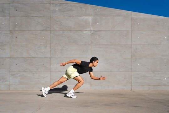 A man in a black shirt and yellow shorts leans forward into a running stance against a gray concrete wall outdoors under a bright blue sky