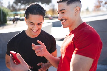 Two men in casual activewear laugh together while holding a smartphone, standing outdoors under the bright sun with trees and a modern background