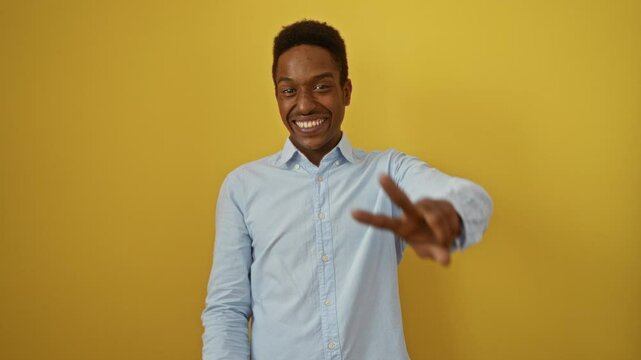 Young african american man standing with a winning smile, wearing shirt, making victory sign with fingers v in cheerful pose over vibrant isolated yellow background