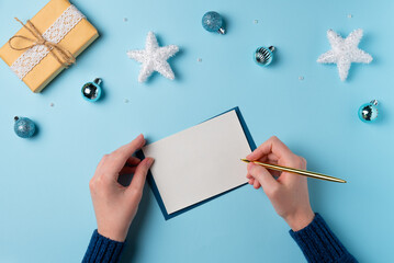 Female hand writing on a blank sheet of paper on a blue background. Christmas concept. Flat lay, top view