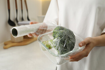 Woman putting plastic food wrap over glass container with broccoli in kitchen, closeup