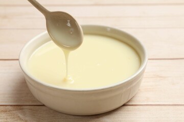Condensed milk flowing down from spoon into bowl on wooden table, closeup