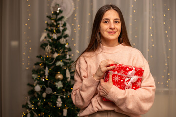 Smiling Woman Holding Christmas Gift in Light Pink Sweater by Decorated Tree, Cozy Holiday Scene with Twinkling Lights, Warm Winter Atmosphere, Festive Spirit, Joyful Christmas and New Year