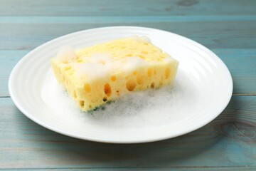 Yellow sponge with foam and plate on light blue wooden table, closeup