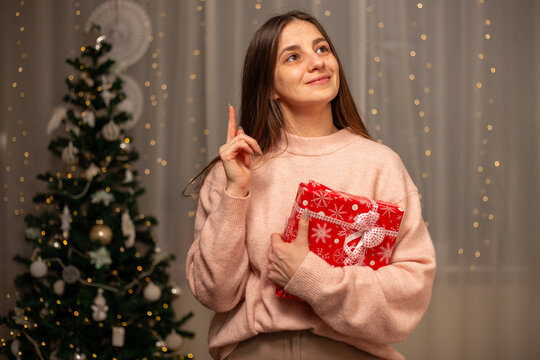 Smiling Woman Holding Christmas Gift in Light Pink Sweater by Decorated Tree, Cozy Holiday Scene with Twinkling Lights, Warm Winter Atmosphere, Festive Spirit, Joyful Christmas and New Year