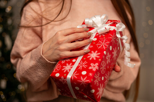 Smiling Woman Holding Christmas Gift in Light Pink Sweater by Decorated Tree, Cozy Holiday Scene with Twinkling Lights, Warm Winter Atmosphere, Festive Spirit, Joyful Christmas and New Year