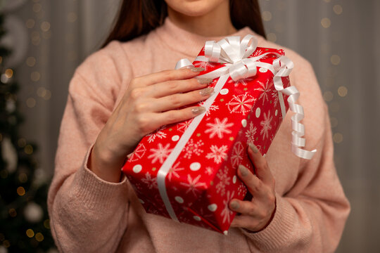 Smiling Woman Holding Christmas Gift in Light Pink Sweater by Decorated Tree, Cozy Holiday Scene with Twinkling Lights, Warm Winter Atmosphere, Festive Spirit, Joyful Christmas and New Year