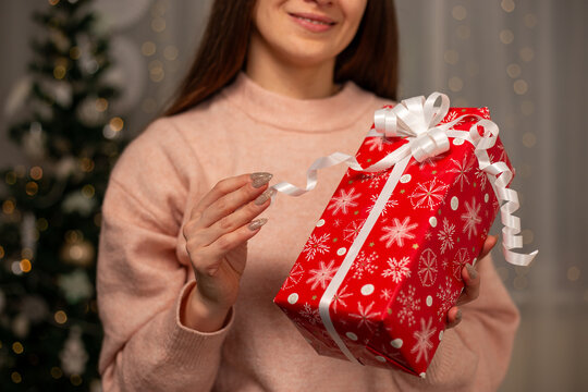 Smiling Woman Holding Christmas Gift in Light Pink Sweater by Decorated Tree, Cozy Holiday Scene with Twinkling Lights, Warm Winter Atmosphere, Festive Spirit, Joyful Christmas and New Year