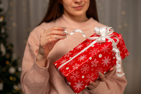 Smiling Woman Holding Christmas Gift in Light Pink Sweater by Decorated Tree, Cozy Holiday Scene with Twinkling Lights, Warm Winter Atmosphere, Festive Spirit, Joyful Christmas and New Year