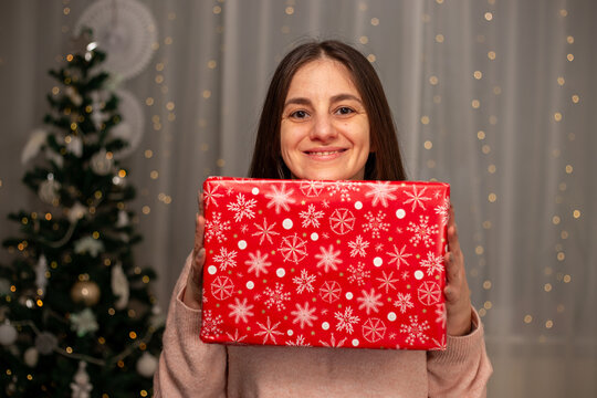 Smiling Woman Holding Christmas Gift in Light Pink Sweater by Decorated Tree, Cozy Holiday Scene with Twinkling Lights, Warm Winter Atmosphere, Festive Spirit, Joyful Christmas and New Year