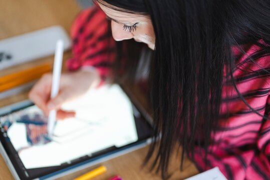 A close-up view of a hand using a stylus to draw intricate patterns on a tablet screen, with colorful pens and a comic book resting on a wooden desk nearby
