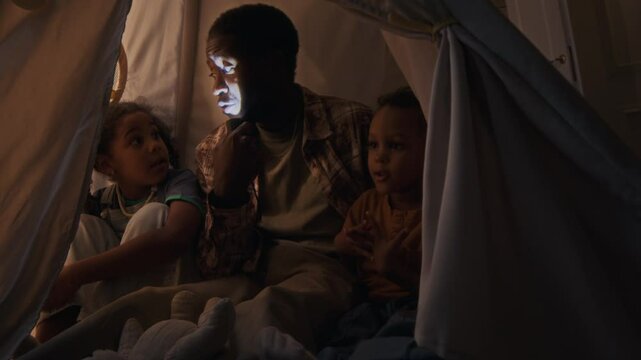 Medium shot of young Black father sitting with little daughter and toddler son inside teepee tent under blanket with flashlight on his face telling scary stories and playing together at night at home
