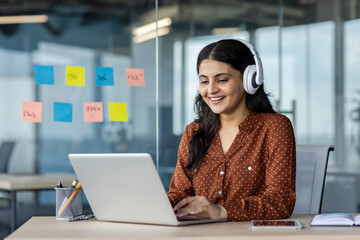 Businesswoman wearing headphones, working on a laptop in a modern office environment, smiling.