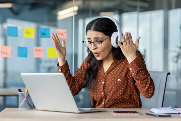 Businesswoman reacts with surprise working on laptop in office space. She wears headphones and...