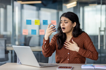 Businesswoman experiencing breathing difficulty in an office using an inhaler as she sits at her...