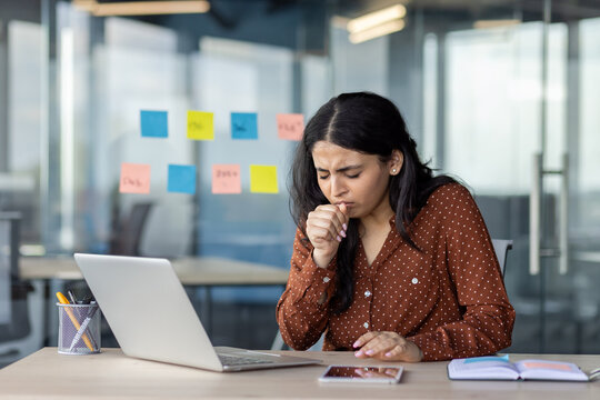 Businesswoman experiencing discomfort and coughing while working on her laptop in a modern office setting, emphasizing concerns about health and workplace conditions during illness or flu season.