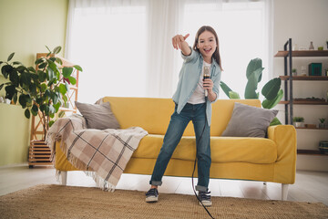 Joyful young girl singing with microphone in living room, wearing casual denim jeans and shirt, enjoying a leisure day indoors at home