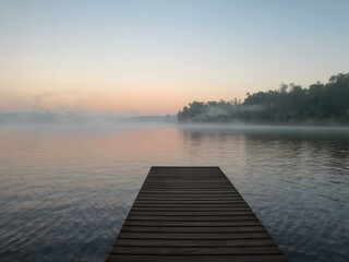 Obraz premium A peaceful evening view of a wooden dock on a misty lake, with soft ripples reflecting a pastel sunset.