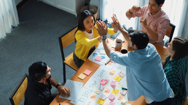 Top view of creative start up team giving high five to encourage their teamwork or successful project while making scrum task board for managing workflow by using marker and sticky notes. Convocation.