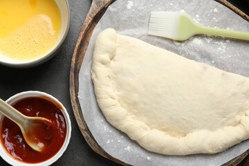 Uncooked calzone pizza, sauce and egg yolk on grey table, flat lay