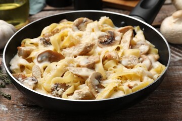 Tasty Alfredo pasta with chicken and mushrooms in frying pan on wooden table, closeup
