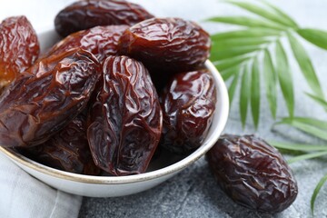Many tasty dried dates in bowl and leaves on gray textured table, closeup