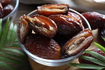 Many tasty dried dates in bowl and leaves on table, closeup