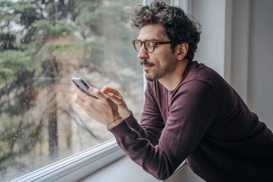 Young handsome middle eastern man scrolling on mobile phone in the morning standing near window.