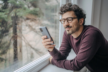 Young handsome middle eastern man scrolling on mobile phone in the morning standing near window.