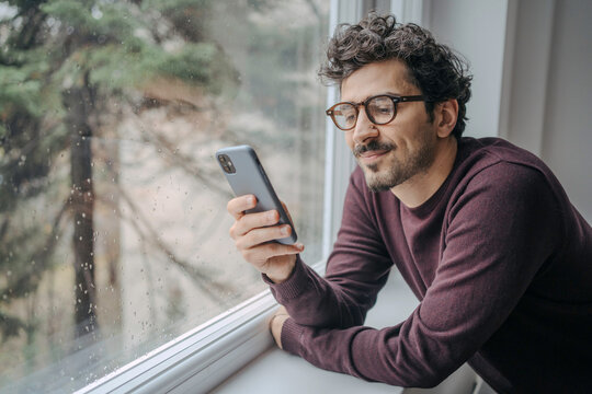 Young handsome middle eastern man scrolling on mobile phone in the morning standing near window.