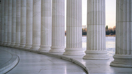 Tall classical white marble columns standing in a row with polished surfaces, marble ground with curved patterns leading to a serene view of water, distant trees, and faintly visible buildings 