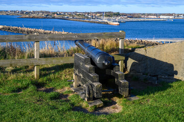 A historic cannon rests on a grassy knoll, providing a scenic view of the tranquil harbor and distant boats on a clear day in a coastal town