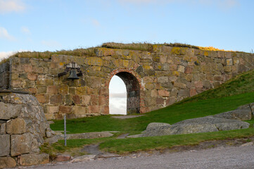 A weathered stone archway with an attached bell stands proudly against a backdrop of rolling green hills. The sun casts a warm glow, enhancing the tranquil scenery