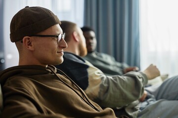 Side view portrait of smiling young man watching TV with friends sitting together on couch at home, copy space
