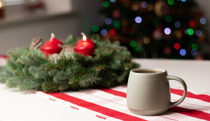 A cup with hot drink on white table with advent wreath and red candles, blurry background with Christmas tree lights