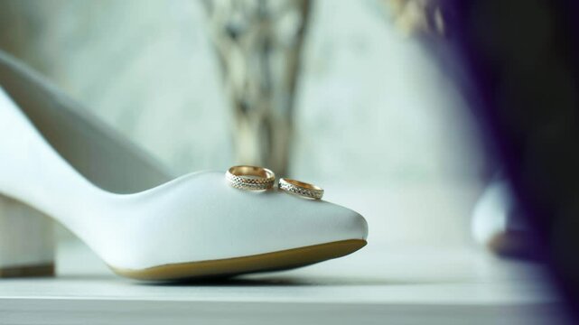 Panning of white female shoe with wedding gold rings on the toe on a light table. Wedding symbols on a light background. Preparing jewellery and shoes for the celebration. No people