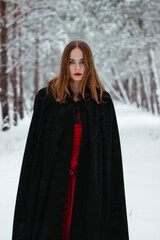 girl in red dress and black cloak walking in snowy forest. woman in medieval dress in winter forest.