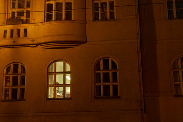 A dark building exterior is presented, featuring a brightly illuminated window at night, which showcases various urban architectural details that add character to the scene