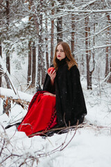 girl in red dress and black cloak walking in snowy forest. woman in medieval dress in winter forest.